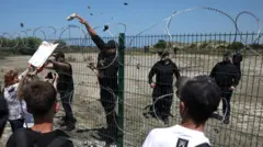 Um arame farpado separa seguranças vestidos de preto e manifestantes com cartazes, como vê um guarda, jogando um projétil de volta por cima da cerca.