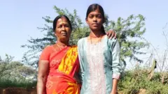 Vasantha veste um sári vermelho com estampa floral amarela, de pé, com a mão no ombro da sua neta Naveena, que veste um top azul, com arbustos ao fundo