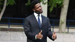 Lil Nas X делает жест большими пальцами двух рук. Он одет в темный костюм, белую рубашку и галстук