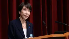 えんじ色の幕を背に演台に向かって話す首相。紺のスーツを着ている