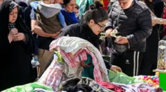 زنی در ۱۷ مارس ۲۰۲۵ در حالی که مردم برای نوروز (نوروز)، سال نو ایرانی، که امسال مصادف با ماه رمضان است، آماده می‌شوند، لباس‌های فروش را در غرفه‌ای خارج از بازار تجریش در مرکز تهران جستجو می‌کند. 