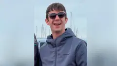 Hayden Davies arimo kumwenyura areba muri 'camera'. 