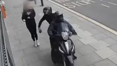 Imagem de CCTV mostrando uma motocicleta preta sendo conduzida por duas pessoas vestidas de preto, roubando um celular de um pedestre que caminhava pela calçada. Uma das pessoas na motocicleta pode ser vista segurando o celular na mão momentos após roubá-lo, enquanto o pedestre recua.