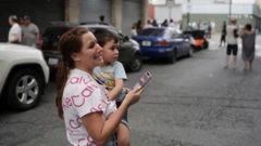 Una mujer con cara de angustia carga a un niño pequeño con una mano mientras sostiene su celular con la otra mano en una calle de Caracas, luego del doble terremoto del 24 de junio.