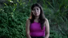 Mulher com blusa rosa com plantas ao fundo.