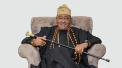 Jide Kosoko joko sori aga pẹlu ọpa lọwọ rẹ, o wọ aṣọ dudu pẹlu fila