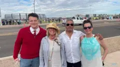 Um grupo de quatro pessoas, dois homens e duas mulheres, sorriem para a câmera, enquanto ao fundo é possível ver um estádio e uma longa fila de pessoas esperando para entrar.