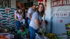 أشخاص يصطفّون داخل متجر صغير للخضار، يختارون منتجاتهم من صناديق مكدّسة؛ امرأة ترتدي قميصاً مخططاً تضع فلفلاً أخضر في كيس في المقدّمة، فيما ينتظر آخرون خلفها. موز وخضروات متنوّعة عند المدخل، ,ويغطي إعلان لفودكا جزءاً من الجدار. 