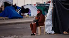 سيدة محجبة ترتدي ملابس باللون الأحمر تجلس على كرسي أمام خيمة باللون الأبيض، وخلفها خيام نازحين أخرى