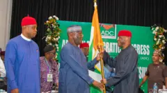 David Mark wey wear black cloth and red cap hold di flag with Atiku Abubakar wey wear blue cloth wit cap wey get designs. ADC Party banner and oda pipo dey for di background