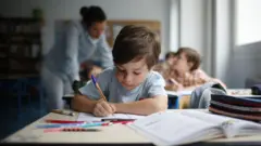 Sınıfta tükenmez kalemle yazı yazan bir çocuk. Arkada kadın öğretmen ve diğer öğrenciler var.