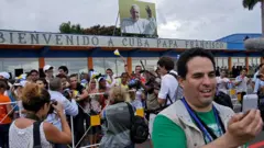 Pessoas em frente a aeroporto, onde se vê foto do Papa Francisco e faixa: Bem-vindo a Cuba, Papa Francisco