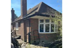 Casa de tijolos de um andar com telhado de inclinação acentuada e duas chaminés. A casa tem janelas com moldura branca, incluindo uma grande janela de sacada na frente. A casa é cercada por vegetação rasteira e há detritos espalhados no chão perto da lateral do prédio. O céu acima está limpo e azul.