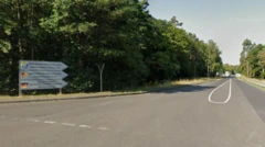 緑に囲まれた車道の交差点に標識が立っている。道の奥には小さな建物と車両を一時停止させるバーのようなものが見える
