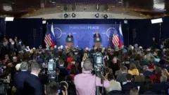 Jornalistas encaram um palco onde Putin e Trump estão de pé em púlpitos. Duas câmeras em tripés aparecem no primeiro plano, operadas por dois homens. Os demais jornalistas estão sentados, agrupados e próximos uns dos outros.