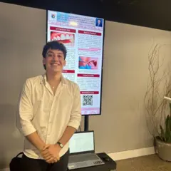 Henry Gaudêncio Almeida veste camisa branca e sorri em frente a um painel com seu trabalho, durante um congresso da área de Odontologia
