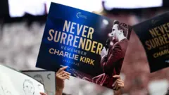 Mãos de mulher segurando um cartaz com os dizeres 'Nunca se renda, lembre-se de Charlie Kirk' e foto de Kirk segurando um microfone.