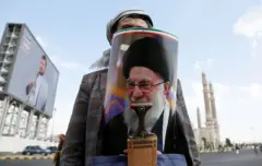 رجل يعلق على إحدى الطرق في صنعاء باليمن ملصقًا صورة المرشد الأعلى الإيراني آية الله علي حسيني خامنئي خلف خنجره في صنعاء باليمن