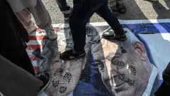 Uma faixa com imagens de Donald Trump e Benjamin Netanyahu estendida no chão é pisoteada por manifestantes