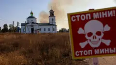 Красная табличка с черепом и надписью по-украински и по-русски: "Стоп мины". На заднем фоне церковь, над которой поднимается дым.