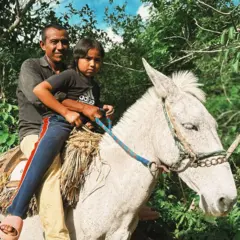 O pai de Ana sorri enquanto ele e a filha andam em um cavalo branco, com sela de palha. Ana veste calças azuis e uma camiseta preta da Adida e seu pai veste uma camisa escura e calças amarelas.