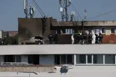 11 funcionarios de Rumania, algunos vestidos de civil con chalecos negros y otros con trajes blancos y cascos inspeccionan la azotea del edificio. En la pared se ve un agujero