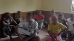 Jovens sentados no chão com os rostos desfocados, encontrados na casa que sofreu a batida da polícia em Makeni, em Serra Leoa