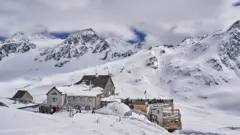 O albergue "Schöne Aussicht Bella Vista". Imagem mostra chalé alpinogame mania cassinodia, rodeado por montanhas nevadas sob céu azul. Esquiadores estão no local, com destaque para a cúpula branca e o terraço com vista panorâmica.