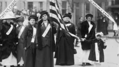 Una foto en blanco y negro de 1909 en el que se ve a mujeres marchando en favor de la prohibición del alcohol en EE.UU. Una en la mitad lleva una bandera estadounidense, la cual es desplegada por una niña- 