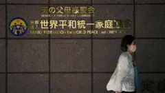 世界平和統一家庭連合(旧統一教会)の文字が張られた壁の前を、マスクをした白い服の女性が歩いている