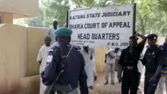 Security operatives guard di main entrance of di Katsina State Sharia Court of Appeal 25 September 2003 for Katsina as d court dey hear Amina Lawal appeal against her death sentence by stoning on top accuse say she commit adultery. 