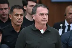 Jair Bolsonaro usando uma camisa preta e com um curativo no pescoço ao lado de policiais