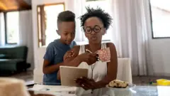 Mulher tomando café da manhã enquanto olha para o notebook e atende um filho ao mesmo tempo