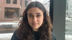 Mia Tretta sits in front of a window in her dorm in Brown University with a street in the background with white snow on the ground. She is wearing a jumper and gold hoop earrings.