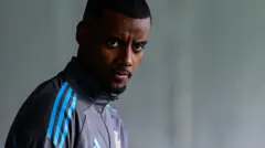 Alexander Isak, vêtu d'un haut d'entraînement noir et bleu à fermeture éclair de Newcastle United, est vu de la poitrine vers le haut, inclinant la tête et regardant directement l'appareil photo, l'arrière-plan étant flou.