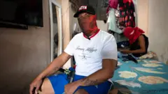 Homem com uma pistola sentado sobre uma cama. Ele veste calção azul, camiseta branca, boné preto e uma balaclava vermelha sobre o rosto