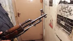 عکسی از یک سلاح کلاشنیکوف و دیواری که در آن مضامین جهادی نوشته شده استږ