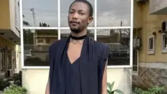 Ezra wear black robe and stand in front of one building surrounded by flowers. E wear choker round im neck and also wear lipstick.