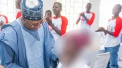 Gómìnà Uba Sani ń rẹrin si ọmọdé kan, àwọn ọkùnrin márùn-ún ń pàtẹ́wọ́