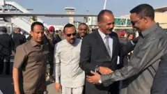 የኤርትራ ፕሬዝዳንት ኢሳያስ አፈወርቂ በ2011 ዓ.ም. አዲስ አበባ ቦሌ ዓለም አቀፍ አየር ማረፊያ ሲደርሱ የተደረገላቸውን አቀባበል ያሳያል። ኢሳያስ በፈገግታ የኢጋድ ዋና ጸሐፊ ወርቅነህ ገበየሁን ሰላም ሲሉ ይታያል። ከአጠገባቸው ጠቅላይ ሚኒስትር ዐቢይ አህመድ እና አምባሳደር ፍጹም አረጋ ቆመዋል።