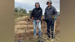 Dave Derby y Peter Derby en un campo arado en septiembre de 2025. Peter está a la izquierda de la imagen, vestido con chaqueta y vaqueros, y Dave a la derecha, con un forro polar y pantalones. Ambos llevan auriculares puestos y detectores de metales en la mano derecha y palas en la izquierda.
