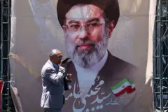 رجل يسير في أحد شوارع طهران، وفي الخلفية صورة للمرشد الأعلى للجمهورية الإيرانية، مجتبى خامنئي يرتدي نظارة نظر وعمامة سوداء ولديه لحية خفيفة تجمع بين اللونين الأسود والأبيض.