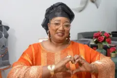 Une femme portant un boubou orange avec des rayures verticales et un foulard noir sur la tête. Elle croise ses doigts en forme de coeur et sourit à la caméra.