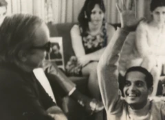 Foto em preto e branco de 1972 mostrando Baden Powell e Vinicius de Moraes em momento descontraído. Vinicius está sentado, de óculos escuros, sorrindo, enquanto Baden Powell, à frente, levanta o braço em gesto animado. Ao fundo, algumas pessoas observam a cena em um ambiente informal.