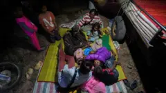 暗がりの中で、地面に敷かれたビニールシートの上に、大人2人と子供たちが数人、身を寄せ合っている。寝袋を羽織っている小さい子供もいる。隣にはリヤカーのようなもののタイヤが見えており、荷台に敷物が重ねてある。