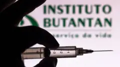 Mão segura seringa diante de logo do Butantant