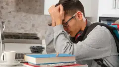 Jovem com os olhos fechados e as mãos na cabeça em sinal de frustração, sentado em frente a livros, cadernos e um computador