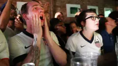 Dois torcedores de futebol dos Estados Unidos reagem durante a partida entre Gana e EUA no bar WoodWork, no bairro do Brooklyn, em Nova York. Um é homem e a outra é mulher. Ambos vestem camisetas da seleção americana de futebol.