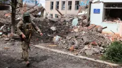 Umusirikare wa Ukraine agenda imbere y'inyubako zasenyutse 