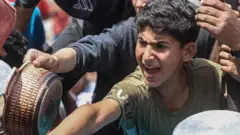 A young boy wearing a dark green tee-shirt and holding a saucepan is leaning forward with his saucepan as he and other people around him wait for food. Behind him is another young boy with a grey t-shirt and he is also holding a pan. Around them other empty sauce pans can be seen. 